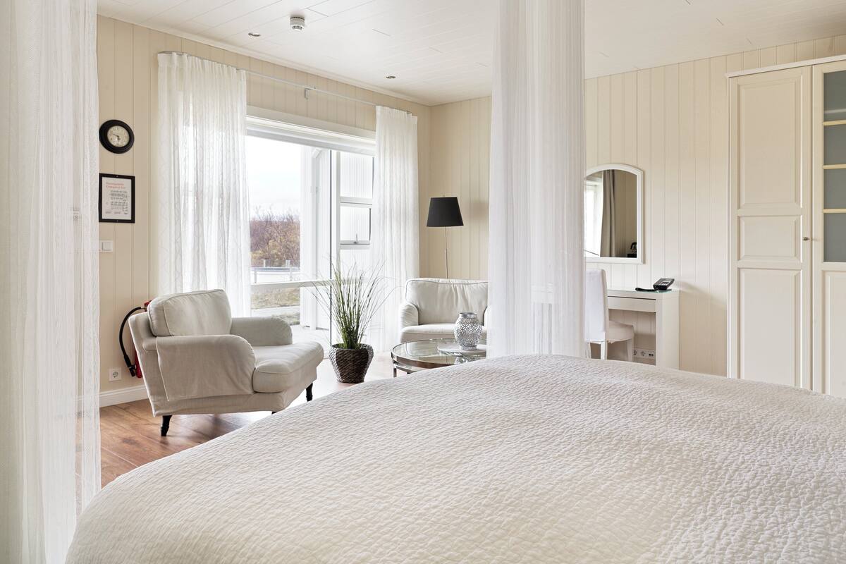 A spacious bedroom with a large bed, two white armchairs, a small table, a potted plant, a mirror, and a window with curtains.