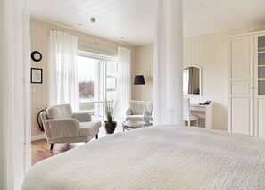 Un dormitorio espacioso con una cama grande, dos sillones blancos, una mesita, una planta en maceta, un espejo y una ventana con cortinas.