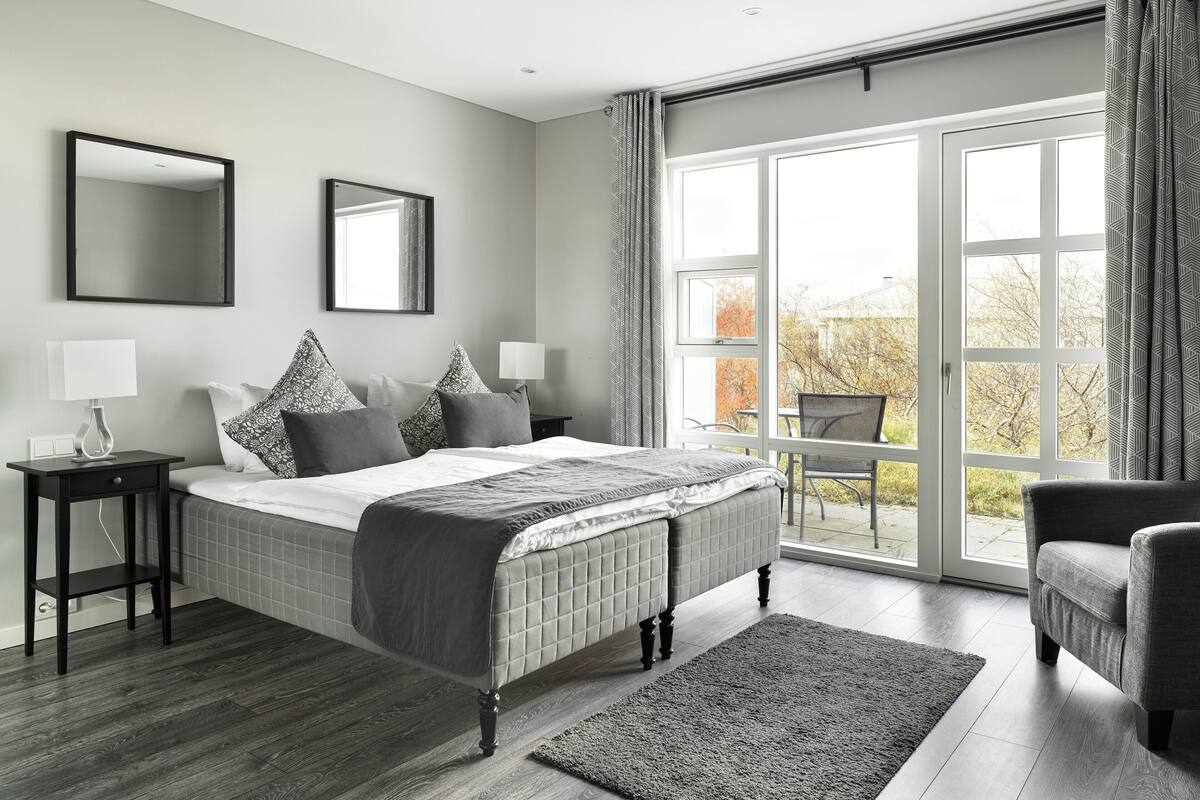 A modern bedroom with a large bed, two bedside tables with lamps, a large window with curtains, and a view of the outdoors.