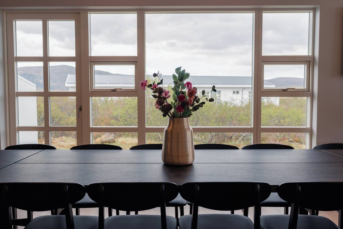 A dining table with a vase of flowers and chairs, set against a large window with a view of a body of water and hills.