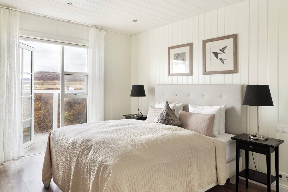 A bedroom with a large bed, two bedside tables, a view through a sliding door, and two framed pictures on the wall.