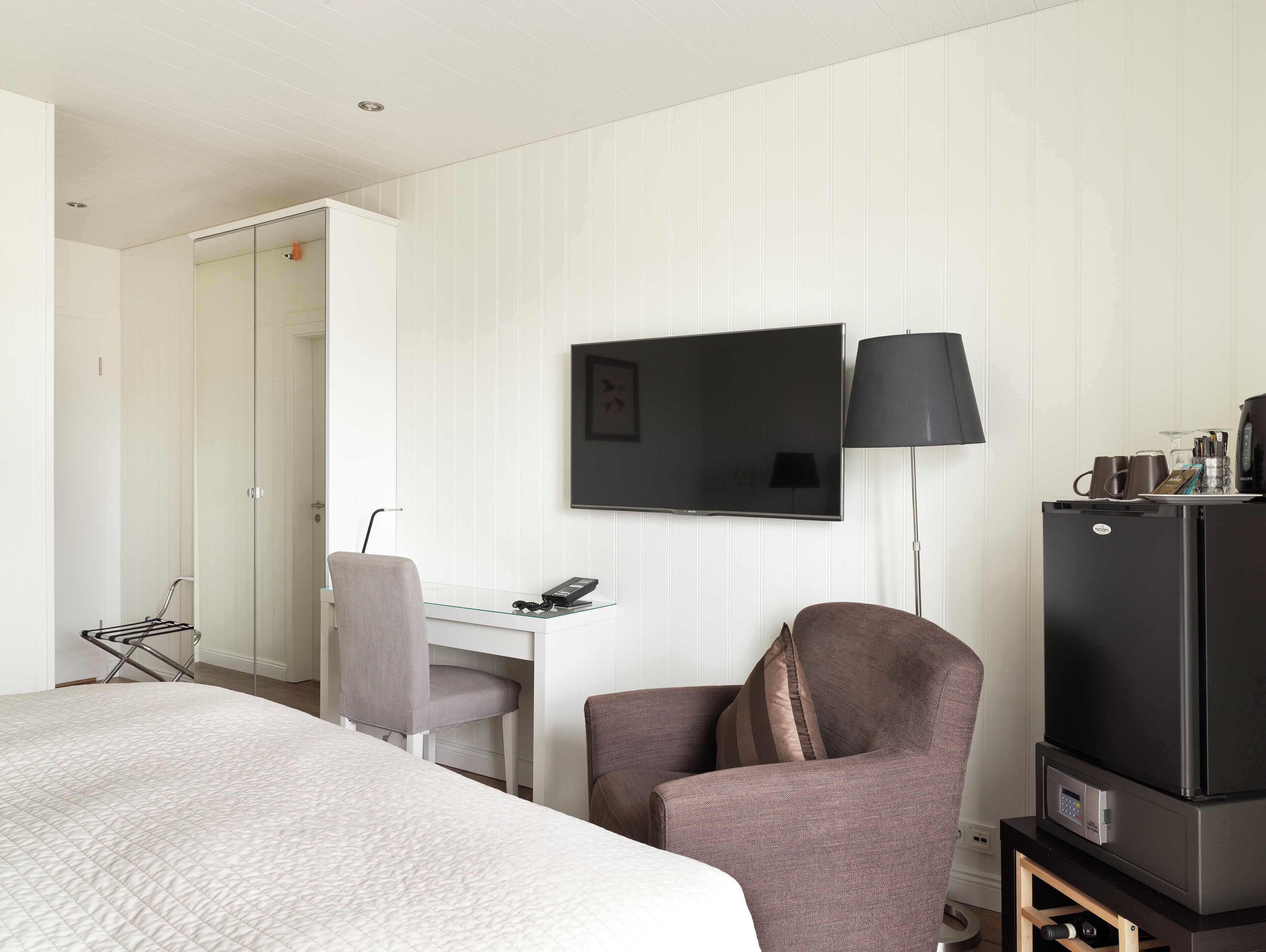 A hotel room with a bed, a desk, a chair, a TV, a lamp, and a refrigerator.