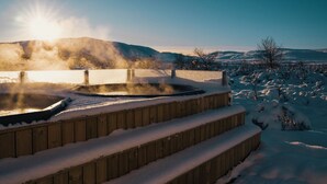 Una bañera de hidromasaje de madera al aire libre rodeada de escalones cubiertos de nieve y paisaje invernal.