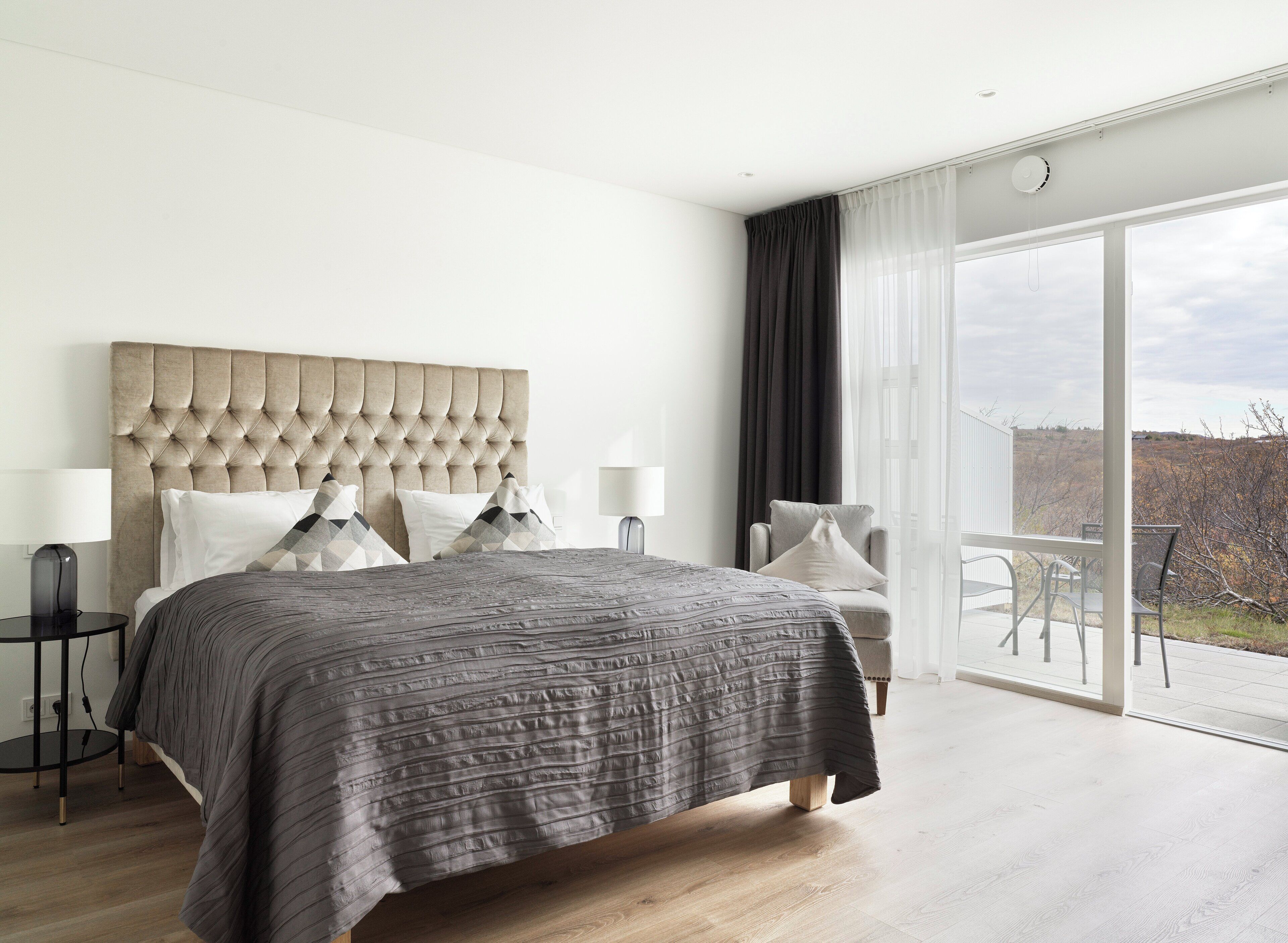 A modern bedroom with a tufted headboard, a large bed with grey bedding, two bedside tables with lamps, a grey armchair, and a balcony with a view of the countryside.