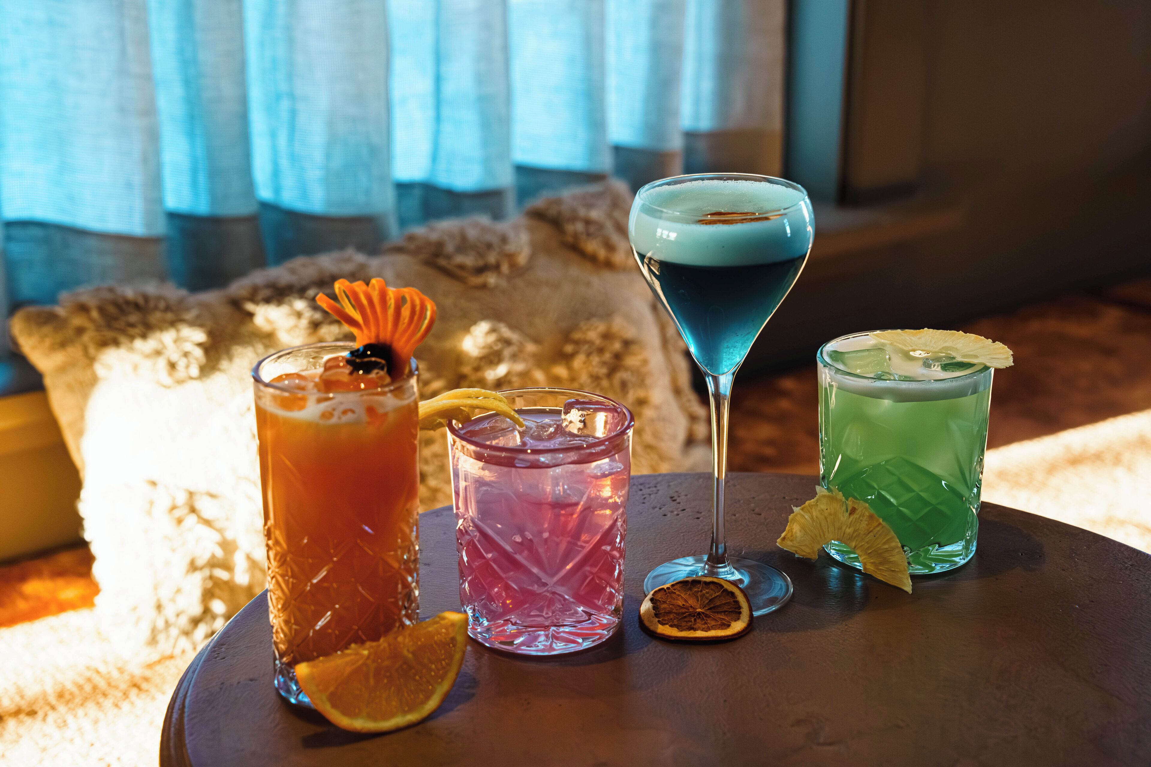 A table with three drinks, each garnished with a slice of fruit, in a room with a window and a textured wall behind.