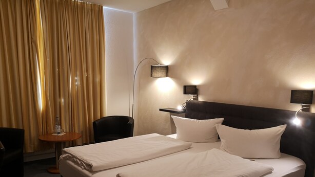 Habitación de hotel con una cama bien hecha, una silla, una mesita con una botella y vasos, y una lámpara de pie.