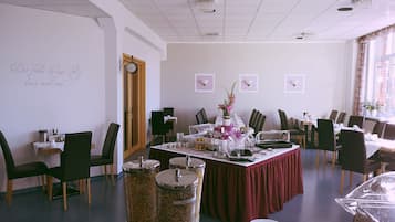 A dining area with a long table set for a meal, surrounded by chairs. There are two trash bins on the floor, a clock on the wall, and a window with curtains.