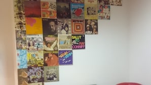 Una habitación con una pared llena de portadas de discos, dos sillas rojas y una mesita con un mantel individual amarillo.