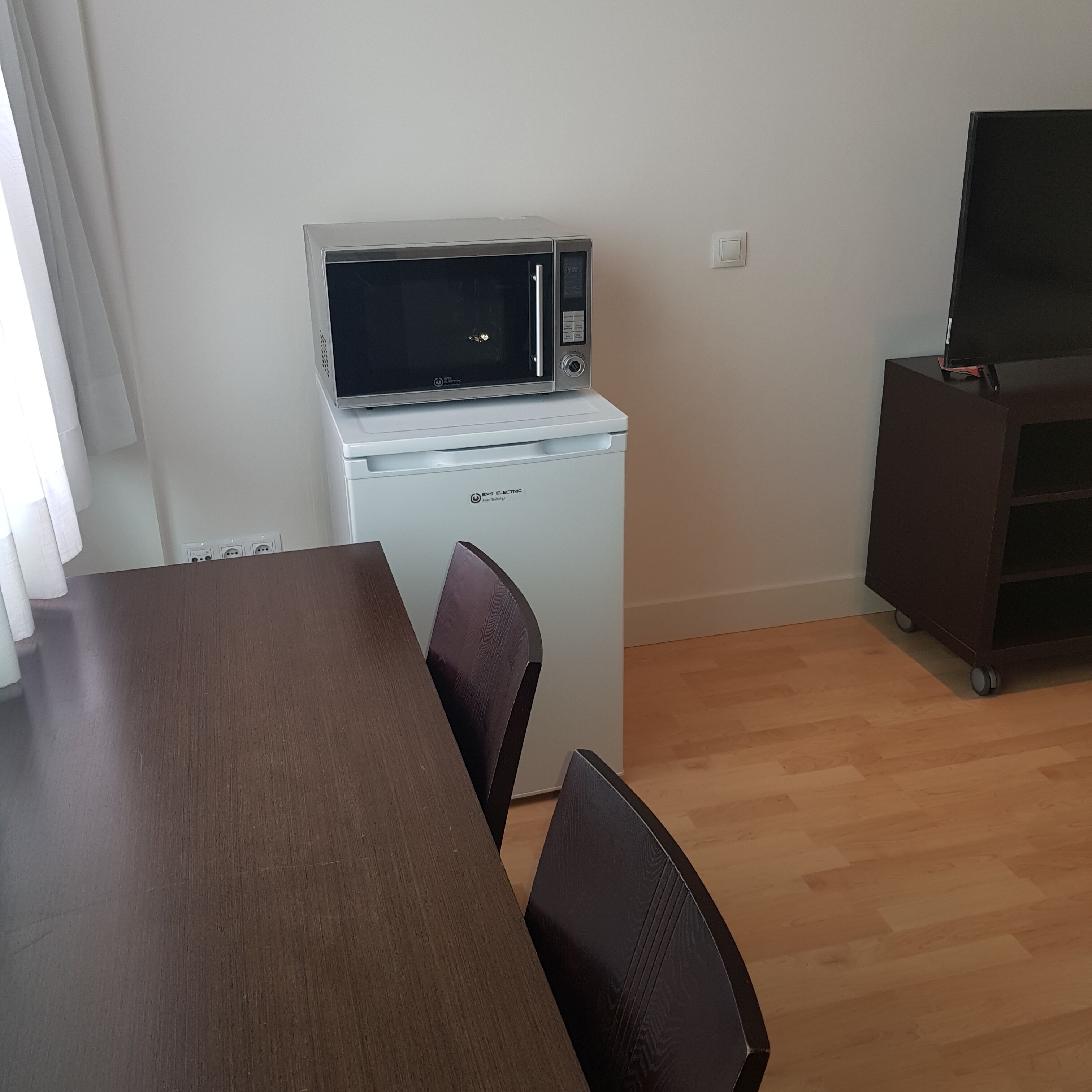 A compact kitchenette with a microwave and a small refrigerator. There is a wooden dining table with chairs and a TV on a stand.