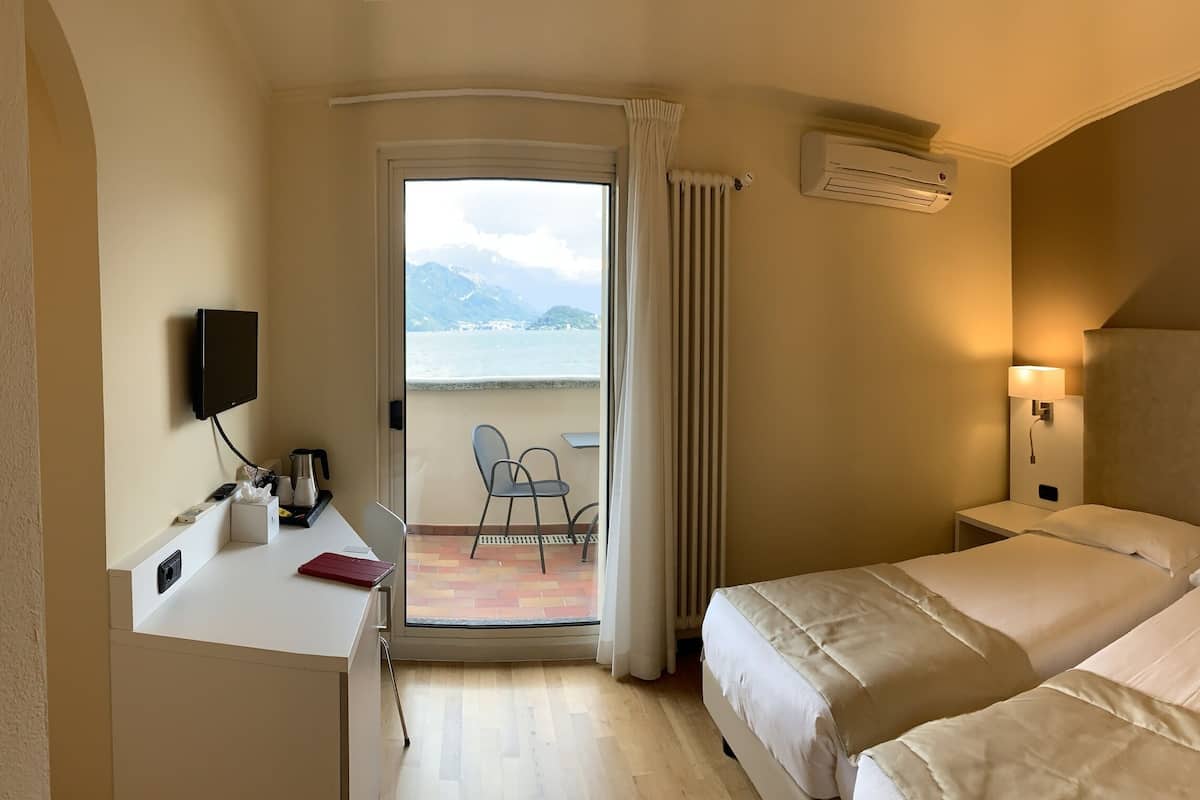 A hotel room with two beds, a desk with a chair, a small table, a TV, and a balcony with a view of the sea and mountains.