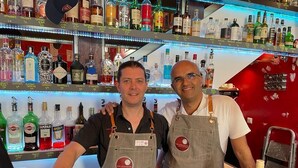 Dos camareros con delantales detrás de una barra repleta de botellas de licores.