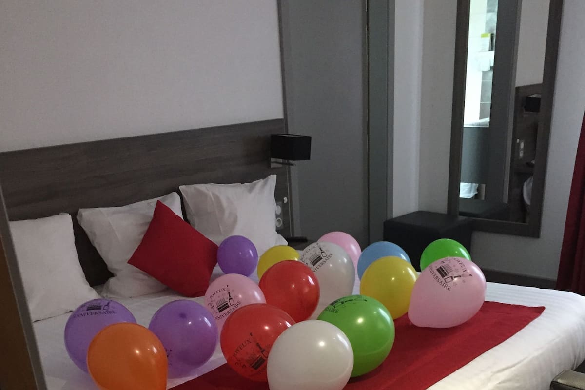 A hotel room with a bed, pillows, and a red blanket. There are colorful balloons on the bed.