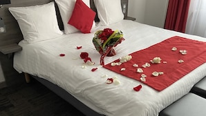Una cama impecablemente arreglada con ropa de cama blanca y detalles rojos, un ramo de flores y pétalos de rosa dispersos en el suelo.