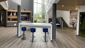 A modern bar area with a white counter, two blue bar stools, and a large window offering a view of the outdoors. - ibis budget Le Mans Universite (Le Mans)