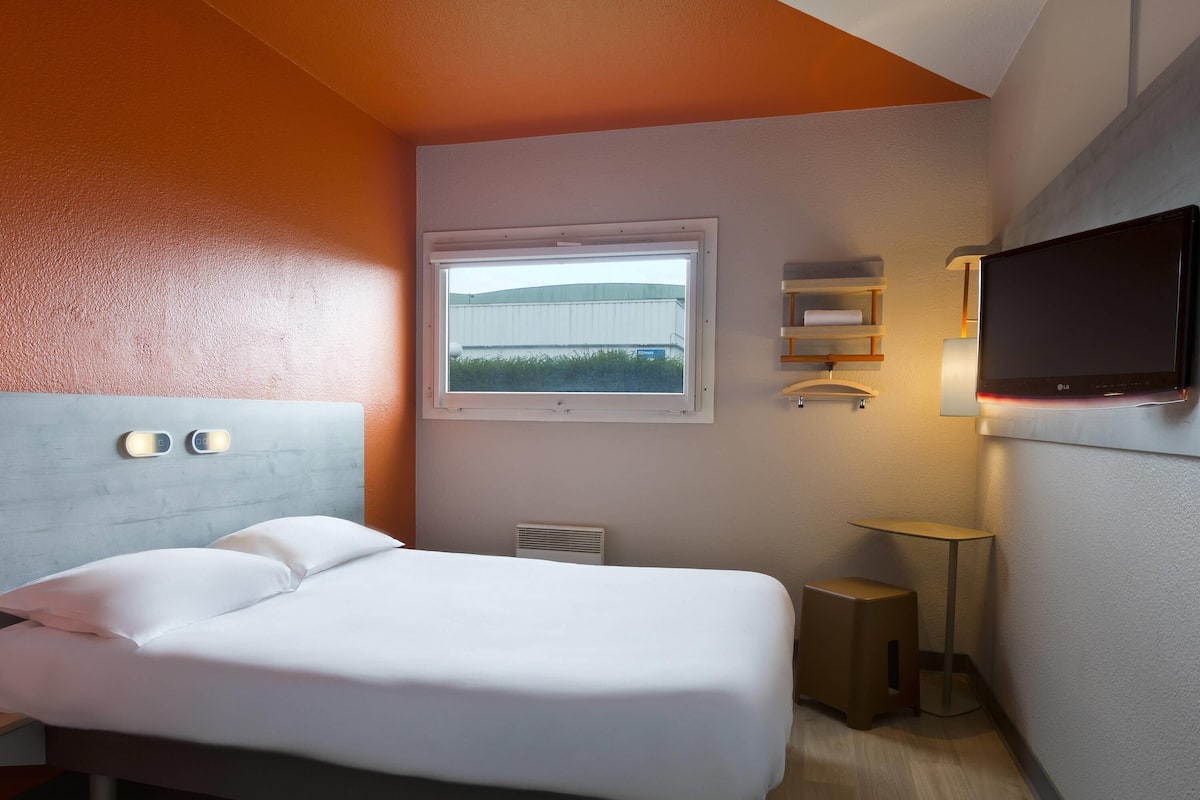 A hotel room with a bed, a TV mounted on the wall, a window with a view, and a bedside table.