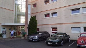 A hotel entrance with a modern design, featuring a reception area, a tall green hedge, and three parked cars. - ibis budget Le Mans Universite (Le Mans)