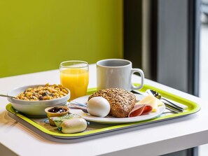 A breakfast tray with cereal, juice, a mug, a bread roll, a boiled egg, a ham slice, and a small container of jam. - ibis budget Le Mans Universite (Le Mans)