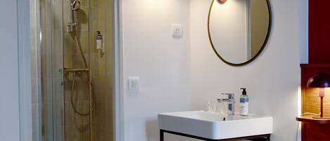A hotel room with a glass-enclosed shower, a white sink with a mirror, and a bed with a colorful blanket.
