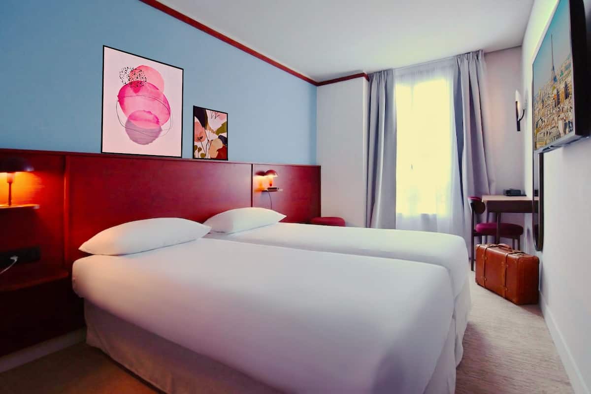 A hotel room with two beds, a desk, a chair, a lamp, and artwork on the walls.