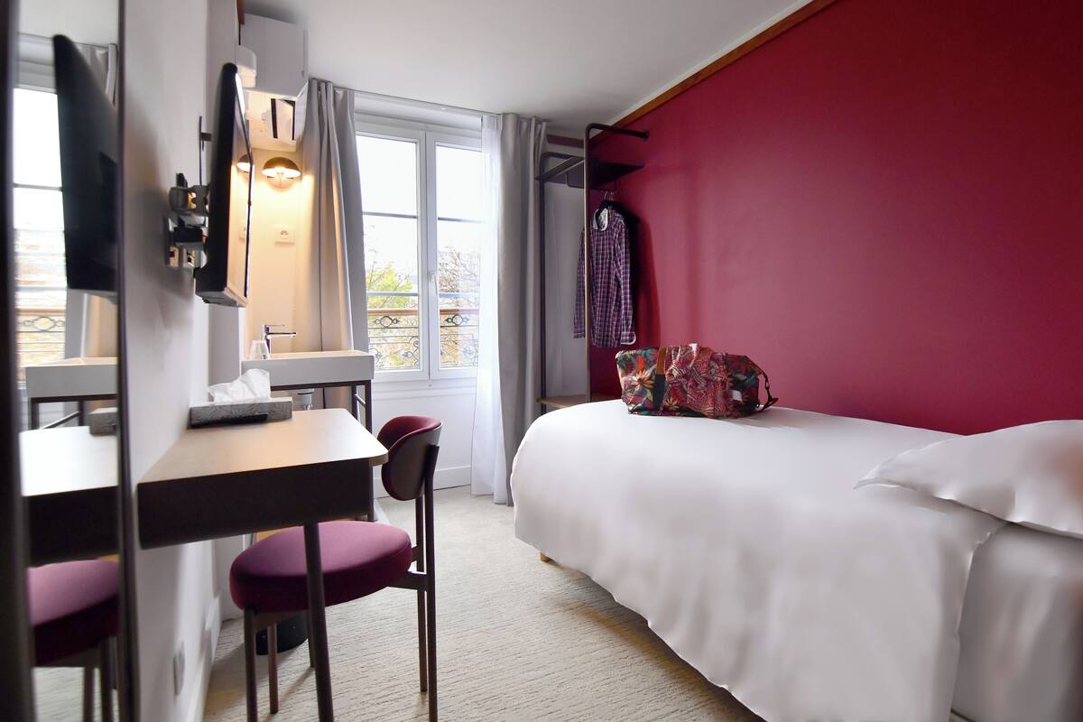 A hotel room with a bed, a desk with a chair, a mirror, and a window with curtains.