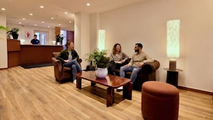 Zona de recepción con piso de madera, un mostrador y tres personas sentadas en sofás.