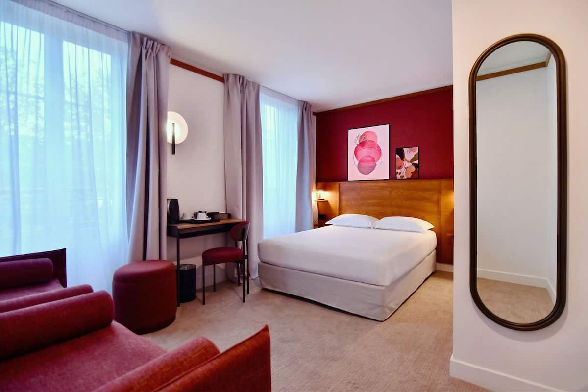A hotel room with a bed, desk, chair, ottoman, and a large mirror.