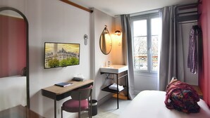 Habitación de hotel con cama, escritorio con silla, baño pequeño, ventana con cortinas y un cuadro en la pared.