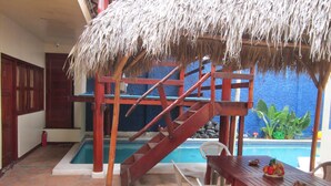 Un área de piscina con una escalera de madera, una mesa con sillas y una estructura de techo de paja.