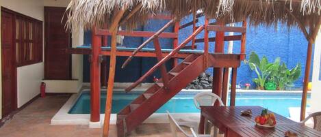 Un área de piscina con una escalera de madera, una mesa con sillas y una estructura de techo de paja.