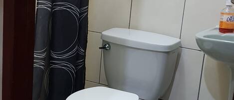 A small bathroom with a toilet, a sink, a shower curtain, a hanging soap dispenser, and a trash bin.