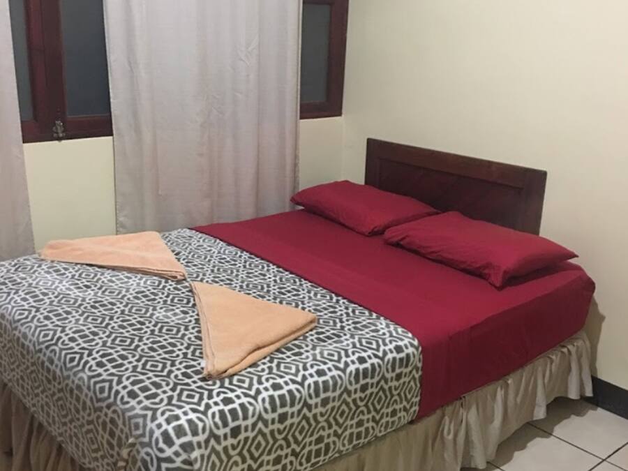 A bed with patterned bedding and a red blanket, a wooden headboard, and a window with curtains.