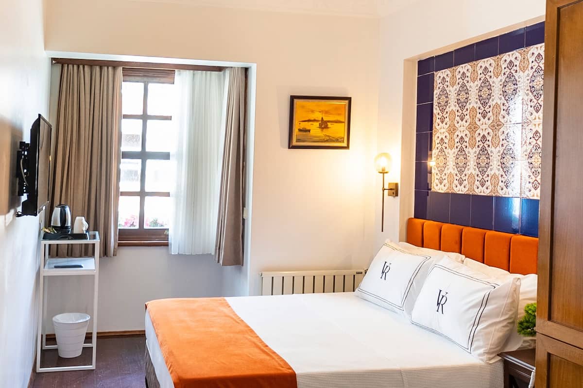 A hotel room with a bed, a nightstand, a radiator, a window with curtains, a wall-mounted lamp, and a decorative wall panel.