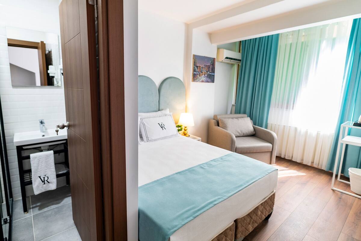 A hotel room with a bed, a chair, a desk, a mirror, a window with curtains, and a bathroom with a towel hanging on the door.