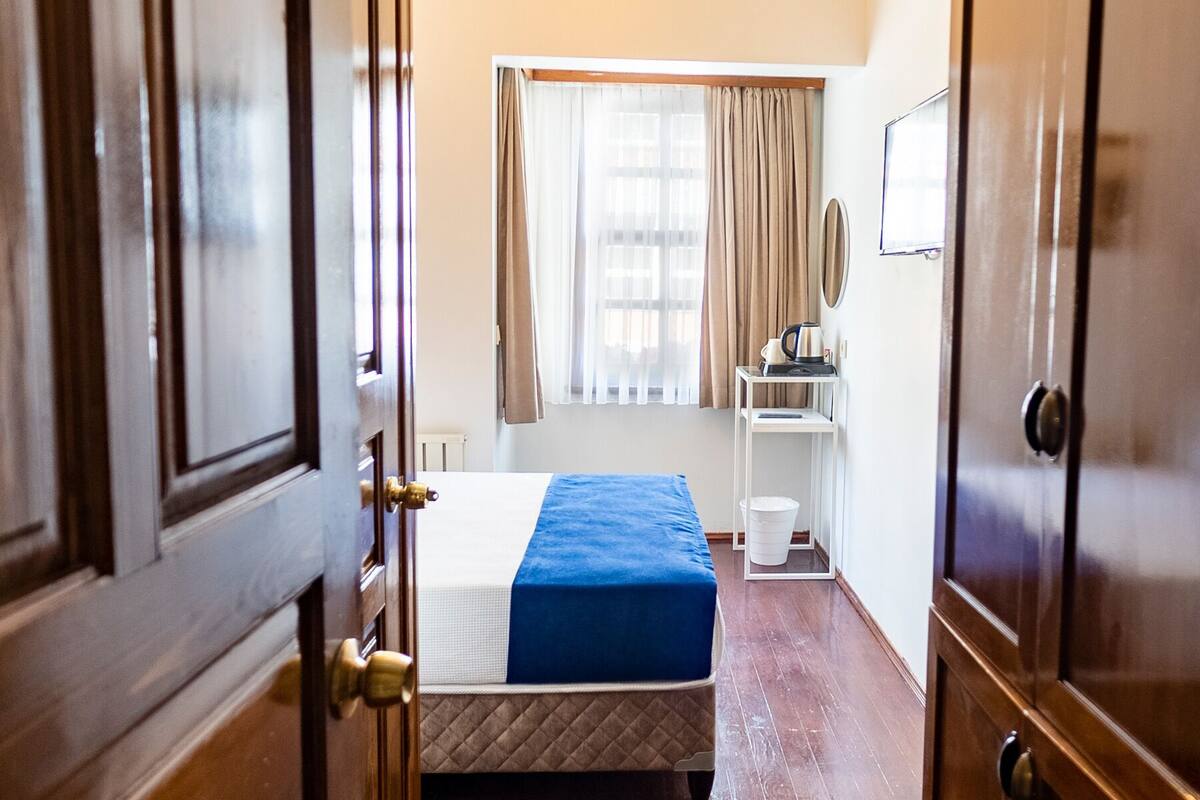 A hotel room with a wooden floor, a bed with a blue bedspread, a window with curtains, a small white table with a lamp, and a wardrobe.