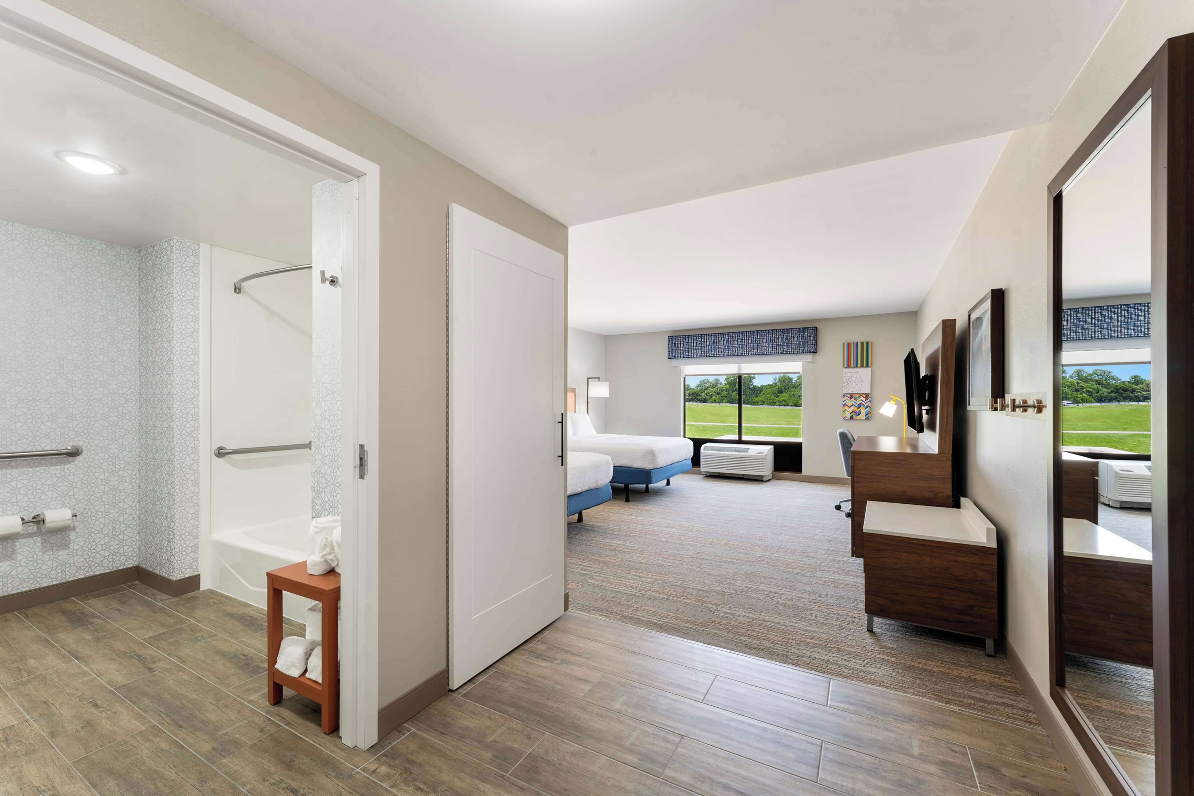 A modern hotel room with a shower, a bathtub, a seating area with a sofa and a chair, and a large window offering a view of the outdoors.
