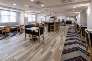 A modern hotel dining area with multiple seating options, including tables and booths, and a central bar area.
