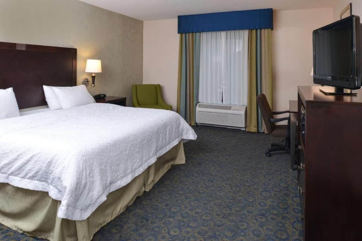 A hotel room with a large bed, a desk with a television, a chair, an air conditioning unit, and a window with curtains.