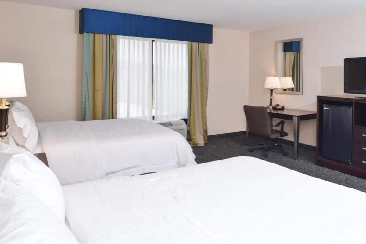 A hotel room with two beds, a desk, a chair, a television, and a window with curtains.