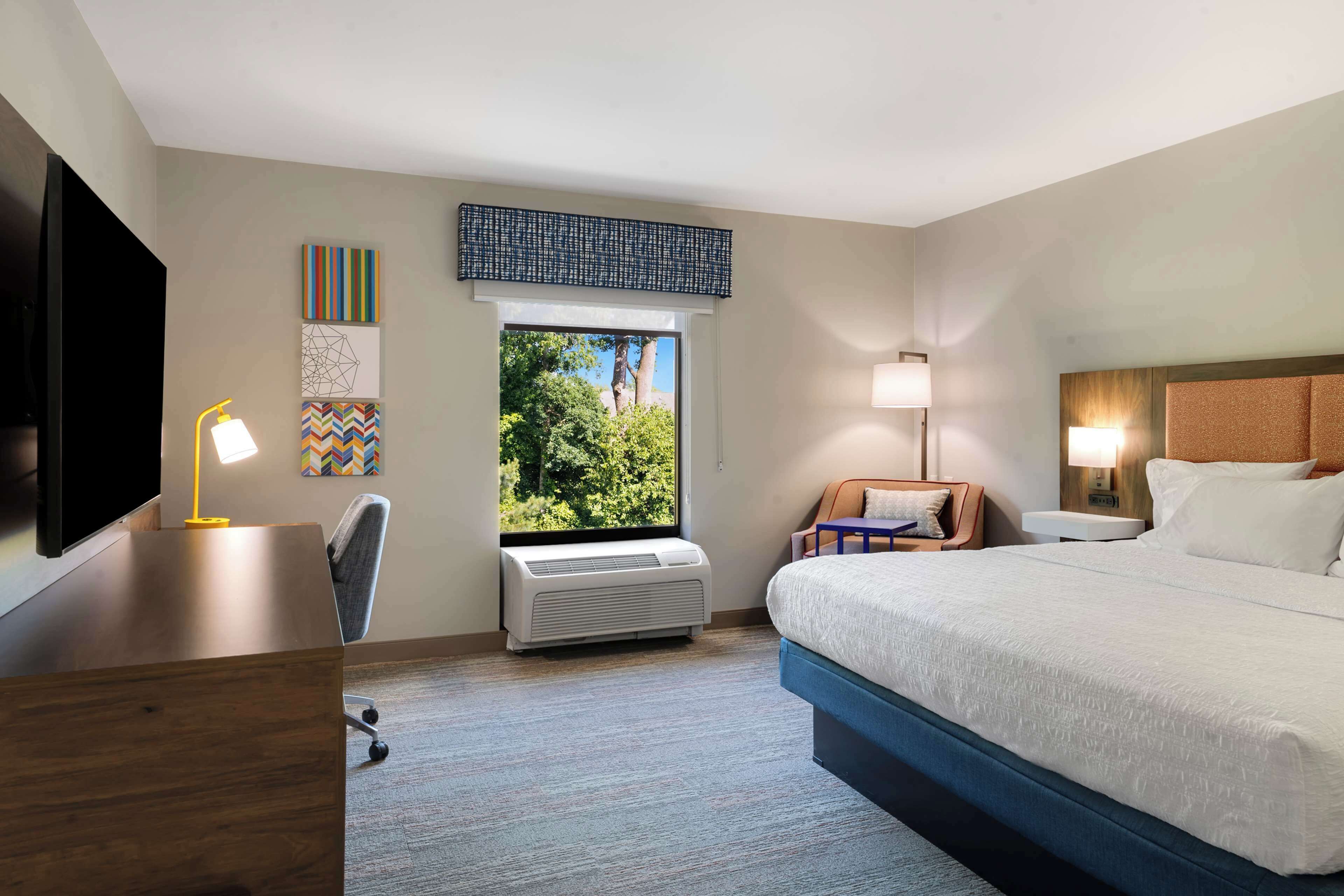 Habitación de hotel con cama, escritorio, silla y un amplio ventanal con vistas a árboles.