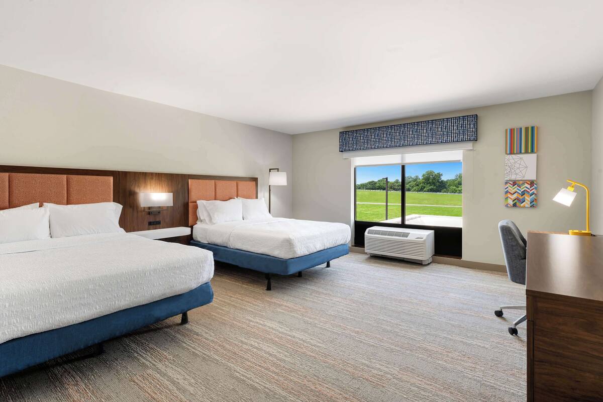 A hotel room with two beds, a desk, a chair, a window with a view of a green field, and artwork on the walls.