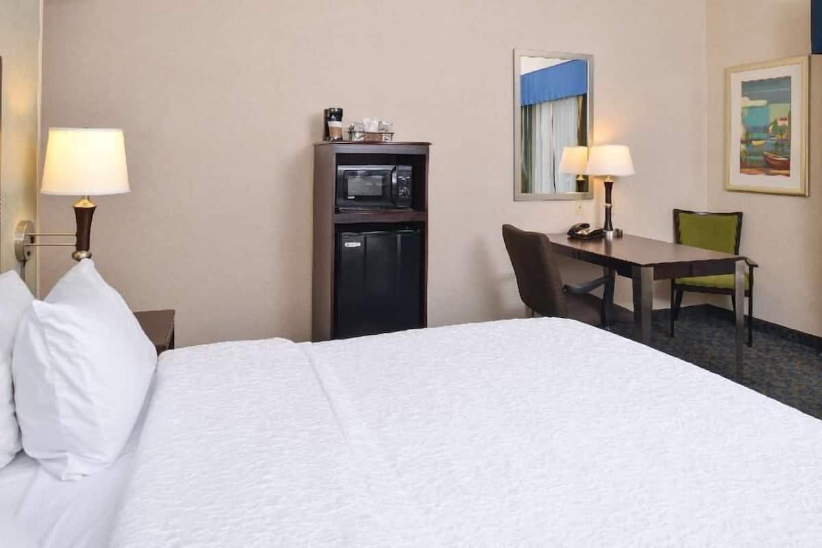 A hotel room with a bed, a desk, a chair, a microwave, a refrigerator, and a lamp.