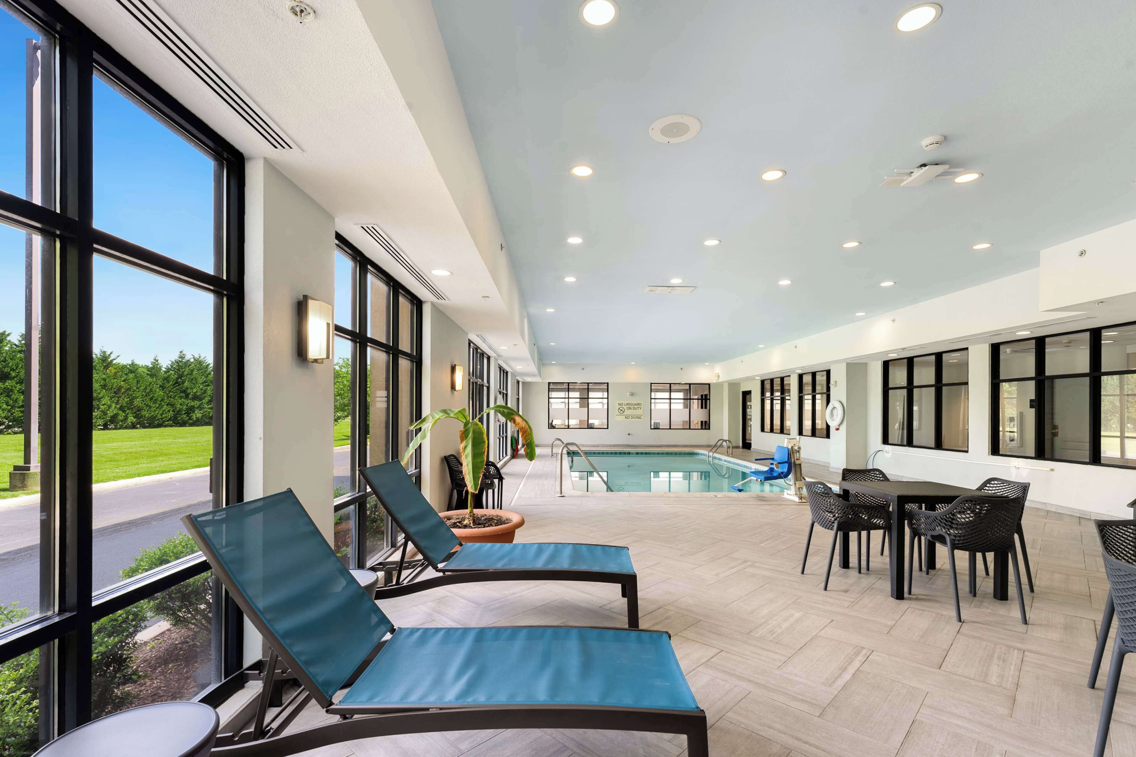A pool area with lounge chairs and a dining table, large windows, and a view of the outdoors.