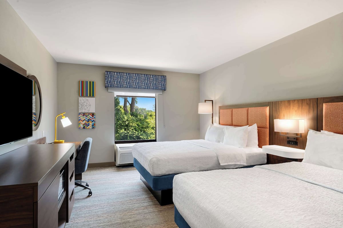 A hotel room with two beds, a desk with a chair, a TV, and a window with a view of trees.
