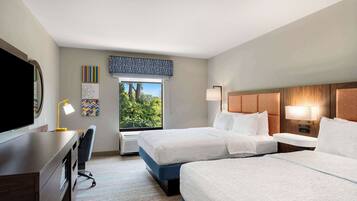 Habitación de hotel con dos camas, un escritorio con silla, televisión y ventana con vista a árboles.