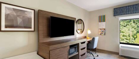 Une chambre d’hôtel avec un bureau en bois, un téléviseur à écran plat, une lampe de bureau, une chaise et une grande fenêtre donnant sur des arbres.