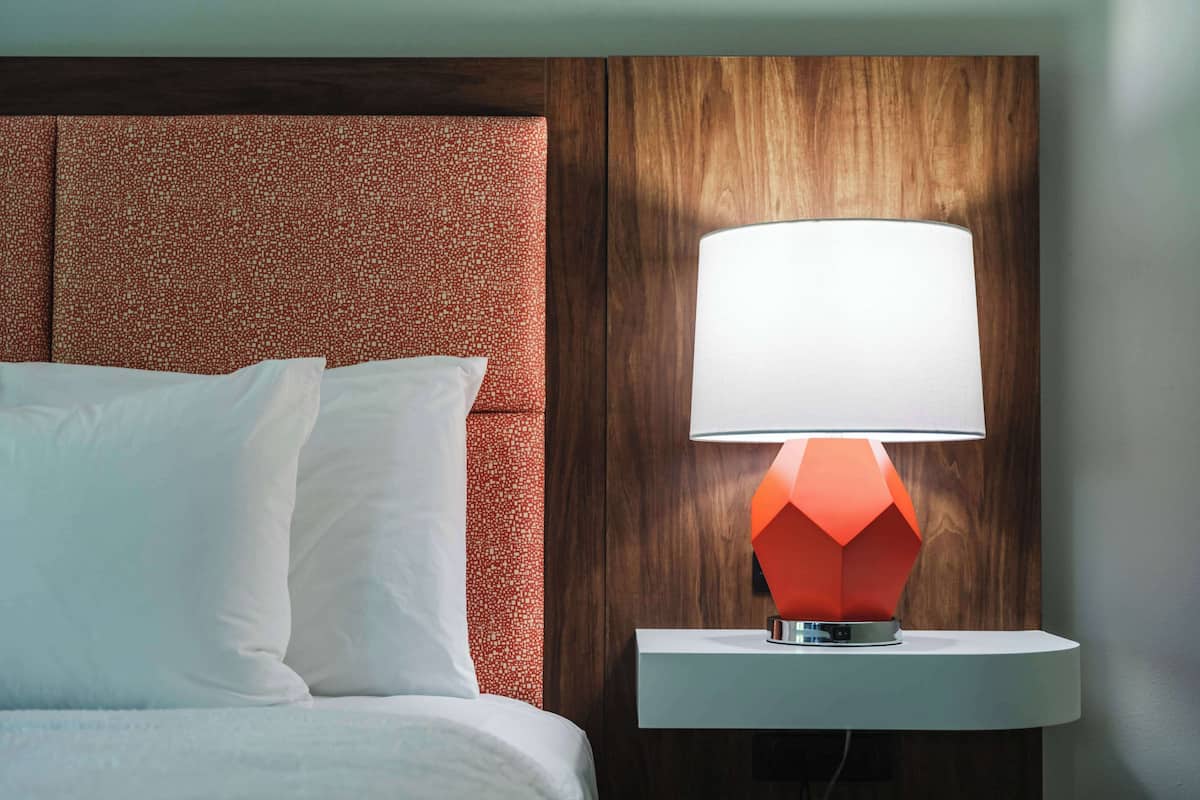 A hotel room with a bed, white pillows, a wooden headboard, and a modern table lamp with a geometric base.