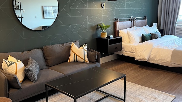 Een moderne slaapkamer met een donkergroene geometrische wand, een ronde spiegel, een bed met wit beddengoed, een donkere bank met decoratieve kussens en een klein bijzettafeltje met een plant.