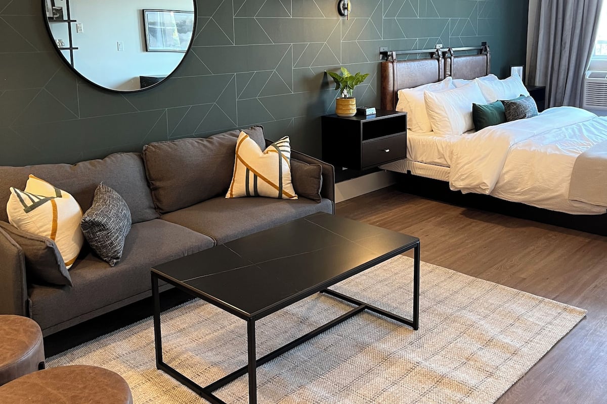 Un dormitorio moderno con pared verde oscuro de diseño geométrico, un espejo redondo, una cama con ropa de cama blanca, un sofá oscuro con cojines estampados y una pequeña mesita de noche con una planta.