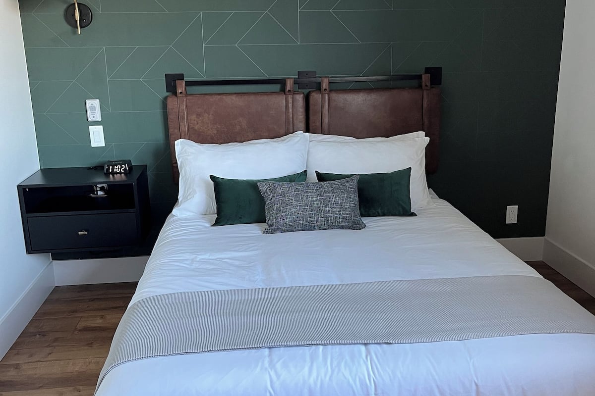 Una cama bien hecha con ropa de cama blanca y verde, un cabecero de madera y una mesita de noche con un reloj.