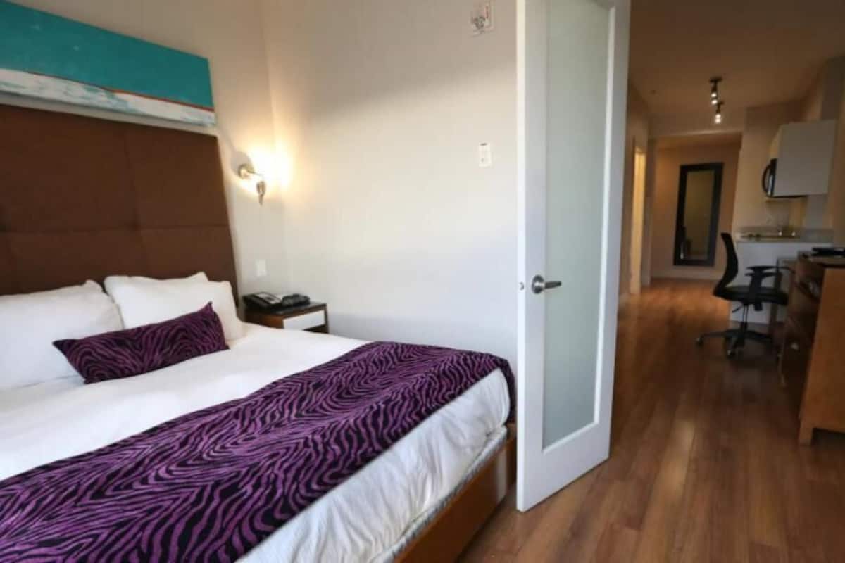 Habitación de hotel con cama, mesita de noche, lámpara y vista al área de la cocina.